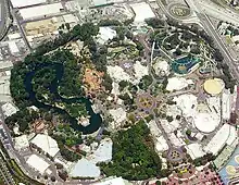 Areal view of a big oval zone with mostly trees and water on the left, buildings and roller coasters on the right.