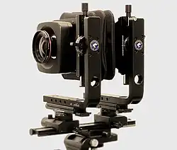 A strange looking device with a lens in front, consisting of bellows in the middle and is held together by two L-brackets on a rail.