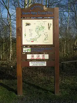 Free standing sign with a map at its center. The caption at the top of the sign reads "Welcome to Severn Valley Country Park". Behind the sign is a grove of trees with enough space between trees to see a blue sky behind them.
