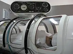 View of the side of a small cylindrical structure with several curved windows and an instrument panel, with a person visible inside
