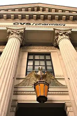 The building's entrance as seen from street level on Eighth Avenue. There is a metal lantern just above the door in the center. The door is flanked by triple-height marble columns that support a triangular pediment. The words "CVS Pharmacy" are mounted below the pediment.