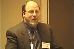 A middle-aged man with balding dirty-blond hair, a white beard, and glasses stands beside a microphone in his grey suit and pattern tie.