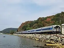 A Metro North train running along the rocky bank of the Hudson. Beyond the train are trees with leave beginning to turn to fall colors.