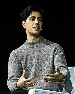 A man with black hair wearing a grey dress speaking into a microphone