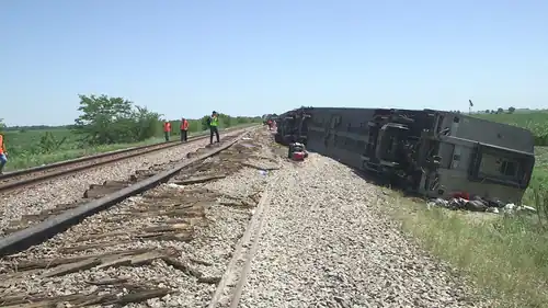 A silver train car turned on its side, with people standing on damaged railroad tracks