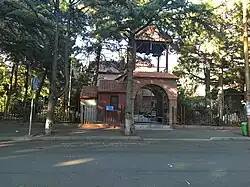 A brick church with a bell tower and an arched entryway, viewed from across a paved road. The church is surrounded by numerous tall trees.
