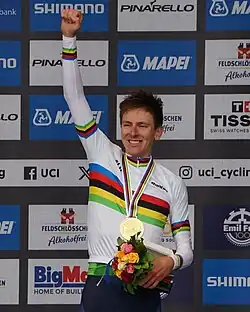 Tadej Pogačar wearing a jersey with rainbow stripes, raising his fist in the air and holding flowers in his other hand.