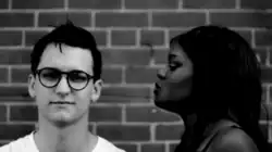 A woman with pigtails talking to a male with glasses who is staring directly into the camera