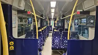 Photo taken inside the train - there are rows of seats on both sides. One side has two seats, and other side has three seats. The seats are blue with multi-coloured spots all over. There are yellow handles for support along the way.