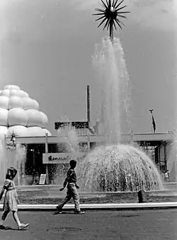 A large fountain in front of a white building. A man and a girl are walking in the foreground.