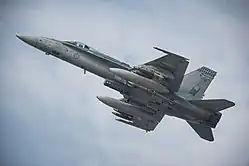 Colour photo of a grey military jet aircraft in flight. The aircraft is armed with missiles and bombs carried below its wings, as well as fuel tanks and a targeting pod.