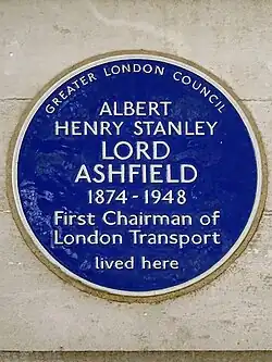 A circular blue ceramic plaque with white raise lettering fixed to a brick wall bears the text "ALBERT HENRY STANLEY LORD ASHFIELD 1874-1948 First Chairman of London Transport lived here".