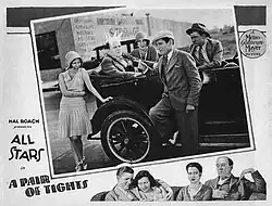 Cinema lobby card. Three men and two women sitting in a car or standing around it. One of the men has four ice cream cones on his head. Both women are laughing. Below the picture is the title of the film and head shots of the four main characters.
