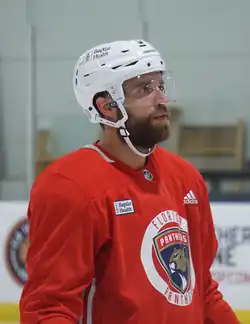 An ice hockey player with a red jersey and white helmet, partially turned away from the camera.