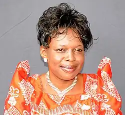 Portrait of a woman in her forties with short, curly hair wearing earrings, a necklace, and an orange dress with a white pattern.
