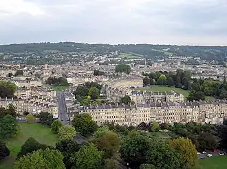 Distance photograph showing rows and crescents of yellow stone buildings. Several trees and hills in the background.