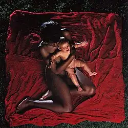 An aerial view of a nude black woman sitting on a red blanket with a white baby held in her arms.