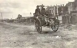 Two men in sombreros riding in a donkey-cart with a line of feet sticking out the back. They are riding down a dirt street away from the camera, with a line of buildings on the right. Dated 5/15/1911.