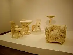 Four cafe chairs and a table, a high table, and an upholstered chair are wrapped in fabric and rope.