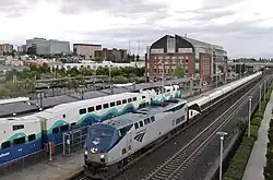 An Amtrak passenger train passes a Sounder train parked on a separate track at a train station with a skyline of mid-rise buildings in the background.