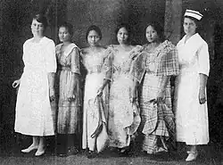 Six women standing in a row indoors. The first woman on the left is white, wearing a white dress. The second woman in Filipina, wearing a western-style dress; the next three women are also Filipinas, wearing "butterfly sleeve" formal dresses; the sixth woman is white, and tall, and wearing a white nurse's uniform and cap.
