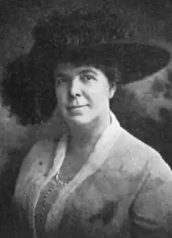 A white woman wearing a large black hat with a brim, and a light-colored suit