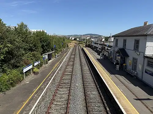 Aerial view of the station taken during the day (with clear blue sky). There are two platforms, with tracks in the centre. On left side, there is the station boundary with a white building on top left corner. The left platform also has a (yellow) shed and a lawn. The right hand side, there is the station office, and parking lot. There is a factory in view. Beyond the platform, there are buildings.