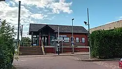 A red brick building with a black roof. It has stairs on the left and a ramp on the right leading up to it. On both sides there are bushes, which slightly obscures the building on the right.