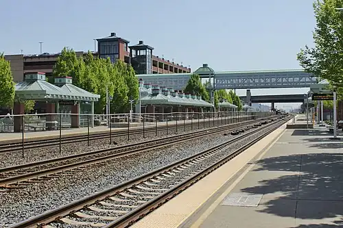 View of a train station with two platforms separated by three railroad tracks that extend into the distance. A large multi-story parking garage, several shelters with canopies, and a covered pedestrian bridge are also in the background.