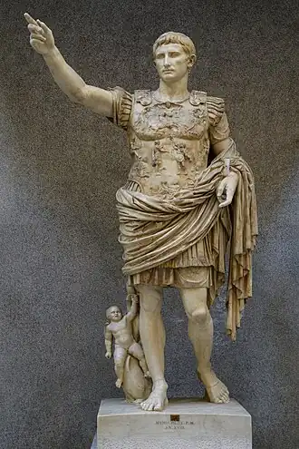 A marble sculpture of the Roman Emperor Augustus. He is pointing up and away from his body with his right hand. He is wearing an intricately carved breastplate depicting his diplomatic skills. At his right foot is a cherub riding a dolphin, a symbol of his family's relationship to the goddess Venus.