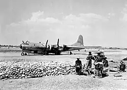 Black and white photograph of the left side of a plane parked on a runway with some Chinese laborers looking on
