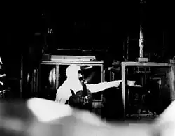 A person in an exposure suit and respirator points to a burnt-out glove box.