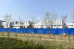 Roadside snapshot of shrubs, grass, and leafless trees. Beyond that, there's an (approximately) 2-meter tall blue fence enclosing a large industrial building. A gardener is tending to the base of one tree on the right.