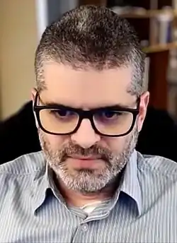 Bernardo Kastrup wearing a light-coloured striped shirt, sitting in a chair, looking downward, seemingly in front of a webcam