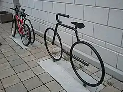 Two bicycle racks shaped like stylized bicycles, in metal with a gloss black finish, each set in a rectangle of concrete within a floor of small ceramic tiles, in front of a white-painted concrete block wall. The one in the background has a mountain bike chained to it.