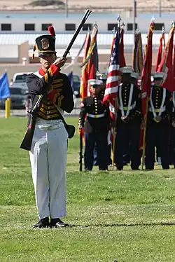 Photo of uniformed man holding a 19th century musket. He wears a replica blue coat and shako from the early 1800s.