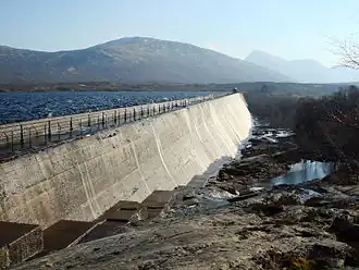 The downstream face of a concrete dam stretching off into the distance, with choppy water in the reservoir behind. To the right is a damp rocky spillway with scrubby trees at the edge of the image. In the distance two mountains are just visible through the haze