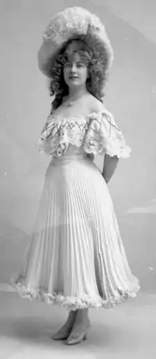 A young white woman, standing, wearing a light-colored pleated and lace-trimmed dress with an off-the-shoulder neckline, and a large hat; her hair is voluminous and falls longer than her shoulders.
