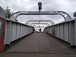 A cast iron footbridge with an upwards gradient. There are regular spans overhead connecting the two sides; the nearest one has a sign that reads "Welcome to Bowes Park"