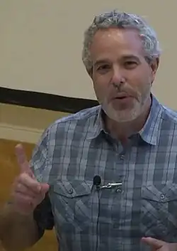 Half-length shot of Frey in a blue shirt and looking towards the viewer, pointing with a finger