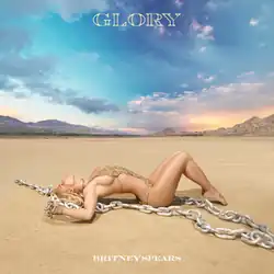 Spears laying in the middle of a desert wearing a gold backless swimsuit with a long chain beside her
