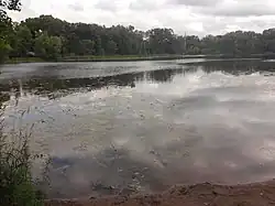 A dark sand beach sits before a dark lake reflecting the cloudy sky above it. Trees and a distant church spire stand on the far shore.