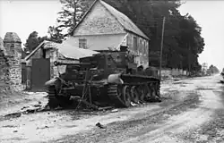 A knocked out tank sits at the side of a road, in front of a two-story house.