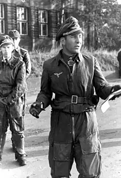 Black-and-white portrait of a man wearing a peaked cap and military flight suit with an Iron Cross displayed at his neck. He is holding a sheet of paper in his left hand and appears to be talking to a larger audience.