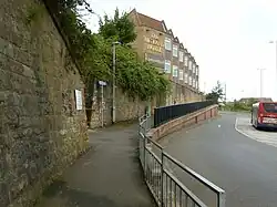 Dark stone viaduct wall to left supporting high level rail track with old railway hotel visible high up having station pedestrian access in centre and bus on a low level roadway exiting bus station on right