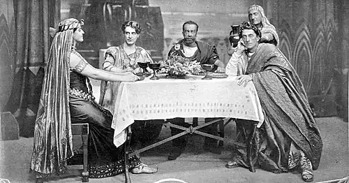 one young woman in Ancient Egyptian dress at a table with three men in Ancient Roman dress, with a servant standing behind them