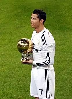 A man in a white shirt poses with a golden ball while standing on football pitch.