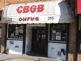 Street view of storefront with doors in the center flanked by windows on both sides. A white awning says "CBGB" in large red letters in a decorative font, and "OMFUG" below that in the same style.