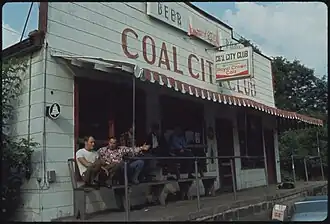 A white building with 'COAL CITY CLUB' in painted in maroon. Some miners sit on benches outside; others "hunker down".