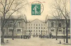 An entrance gate between two gatehouses, opening onto a vast courtyard, the latter enclosed in the background by a four-storey building.